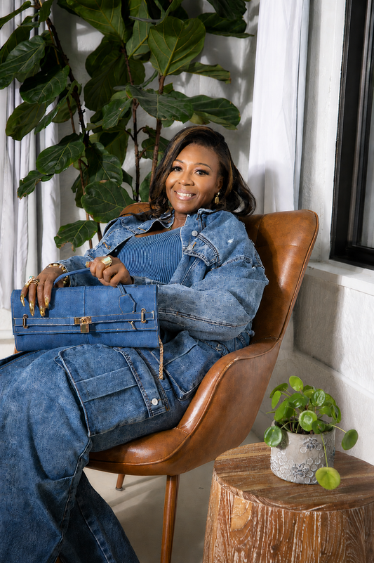 A woman wearing a blue denim jumpsuit and matching blue handbag sits in a brown leather chair in a bright room with indoor plants.